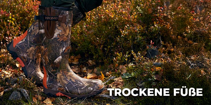 Person trägt camouflagefarbene Gummistiefel der Marke Viking und steht in feuchtem, herbstlichem Gelände; daneben steht der Schriftzug ‚Trockene Füße‘.